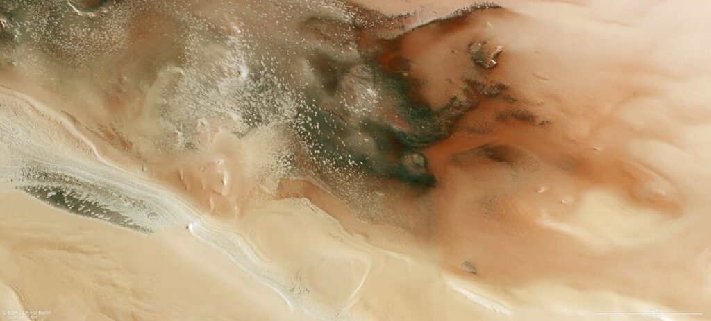 mars express Immagine della superficie di Marte. Una serie di strati chiari e scuri esposti sul fianco di una ripida scogliera si snoda dal centro a sinistra dell’immagine fino al centro in basso. La parte inferiore sinistra e destra dell’immagine è liscia e ondulata. Al centro dell’immagine domina un motivo a chiazze di poligoni scuri di forma irregolare, i cui bordi sono ricchi di ghiaccio brillante. Crediti: Esa/Dlr/Fu Berlin